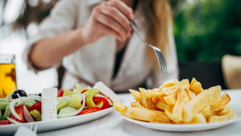 Grande plano de uma mulher a comer uma salada e um prato de batatas fritas - alusão a erros nas dietas
